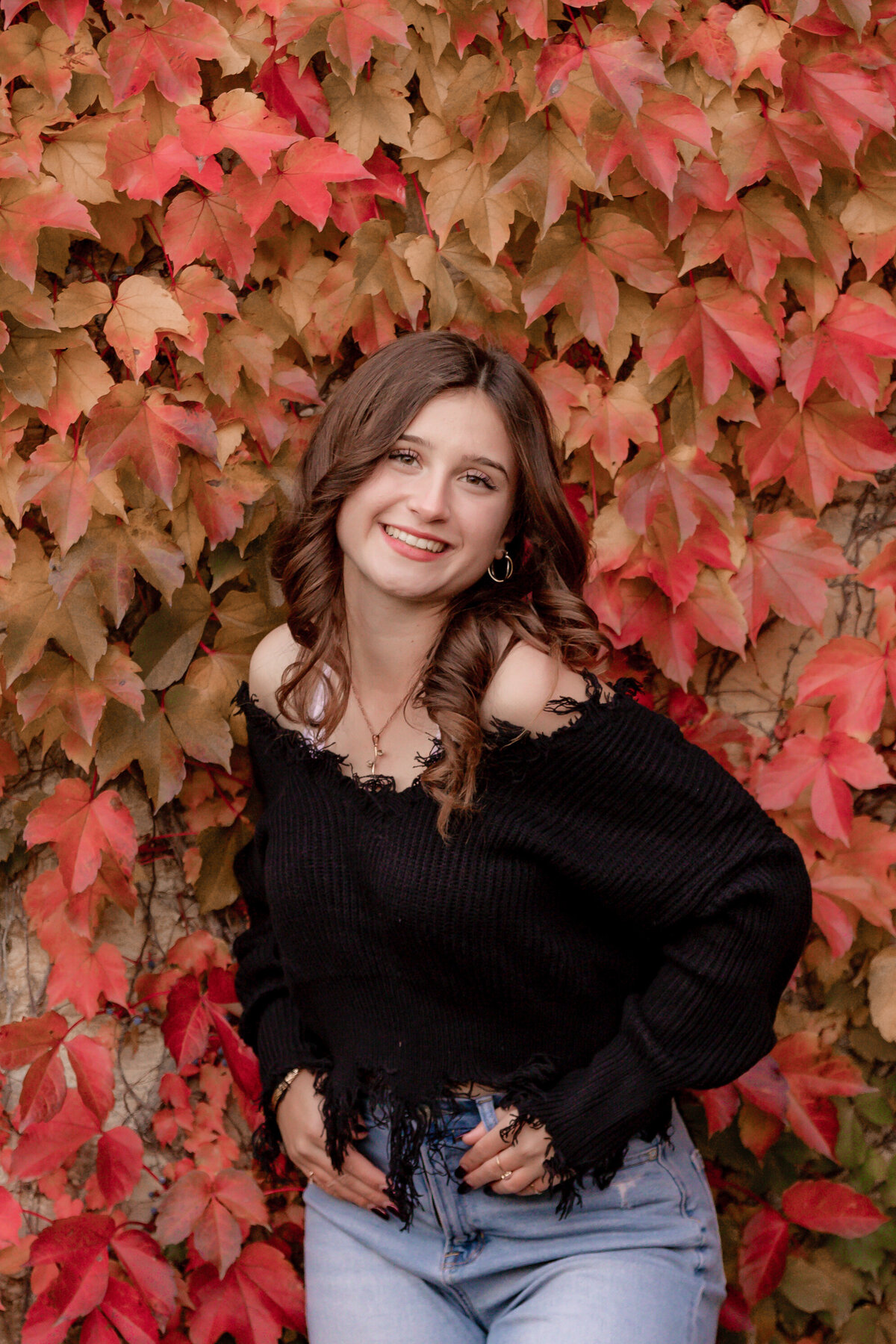 A high school girl in a black sweater poses in front of an orange ivy wall.