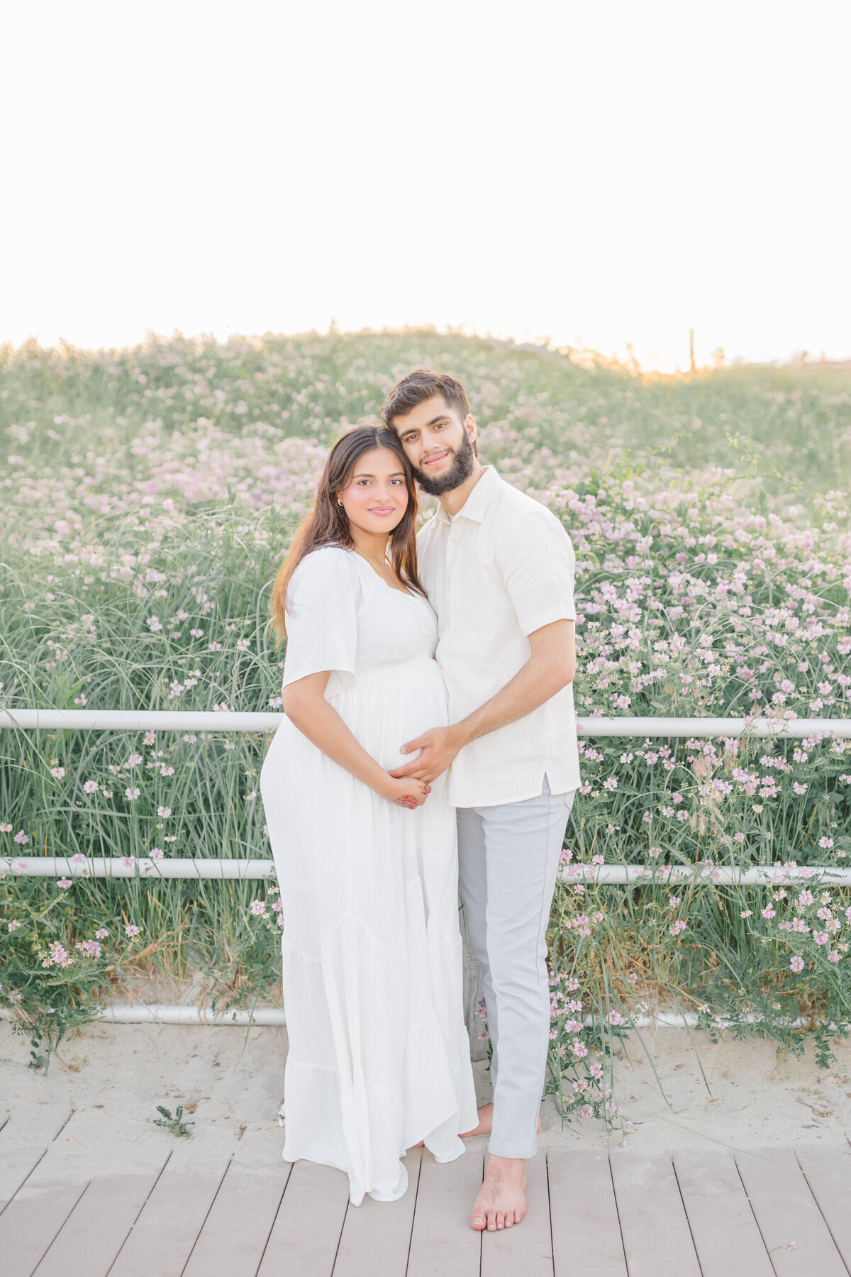 A glowing pregnant woman in a flowing white dress stands on a boardwalk, cradling her baby bump as her partner embraces her with a loving smile. Behind them, soft pink wildflowers sway in the warm evening light, capturing a moment of pure anticipation and joy