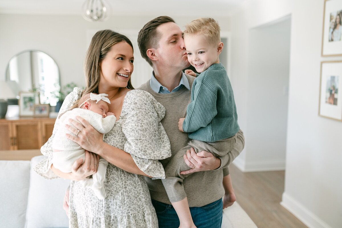 Mom holds newborn daughter on her chest while smiling over at her husband who is holding her son. They are standing in the living room