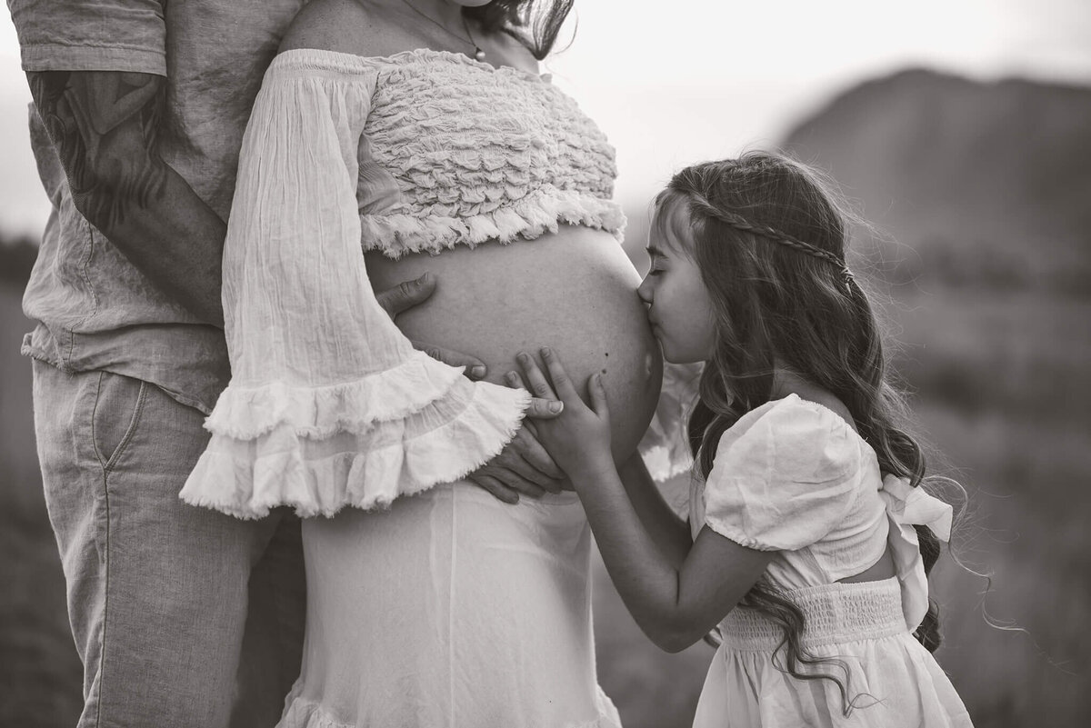 A black and white portrait by Brittany Danielle Photography of a young girl crouching down to kiss pregnant moms belly. Dad stands behind mom embracing her,