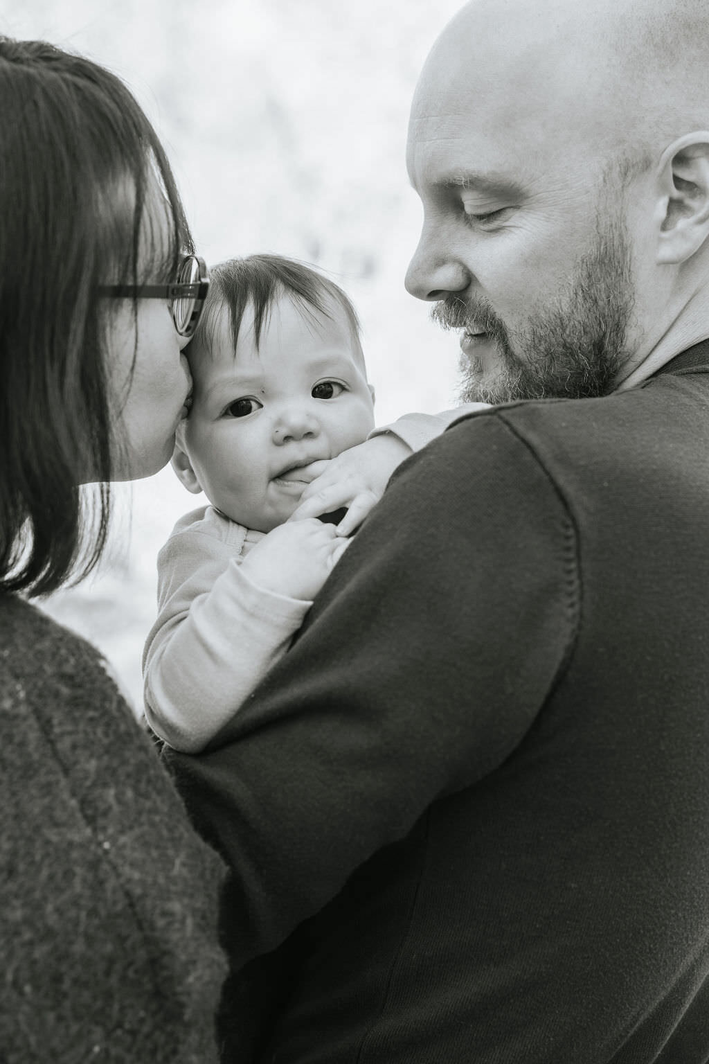 A parent holding their child while their partner kisses the baby's head.