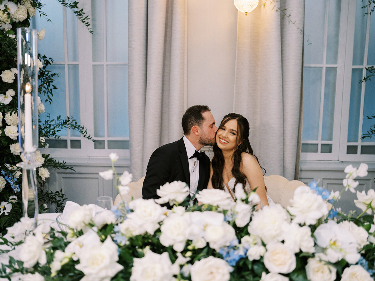 A man in a black suit kisses a woman in a white dress on the cheek. They sit together behind a table adorned with white flowers, under a softly lit chandelier, celebrating their Fairmont Palliser Wedding Calgary.