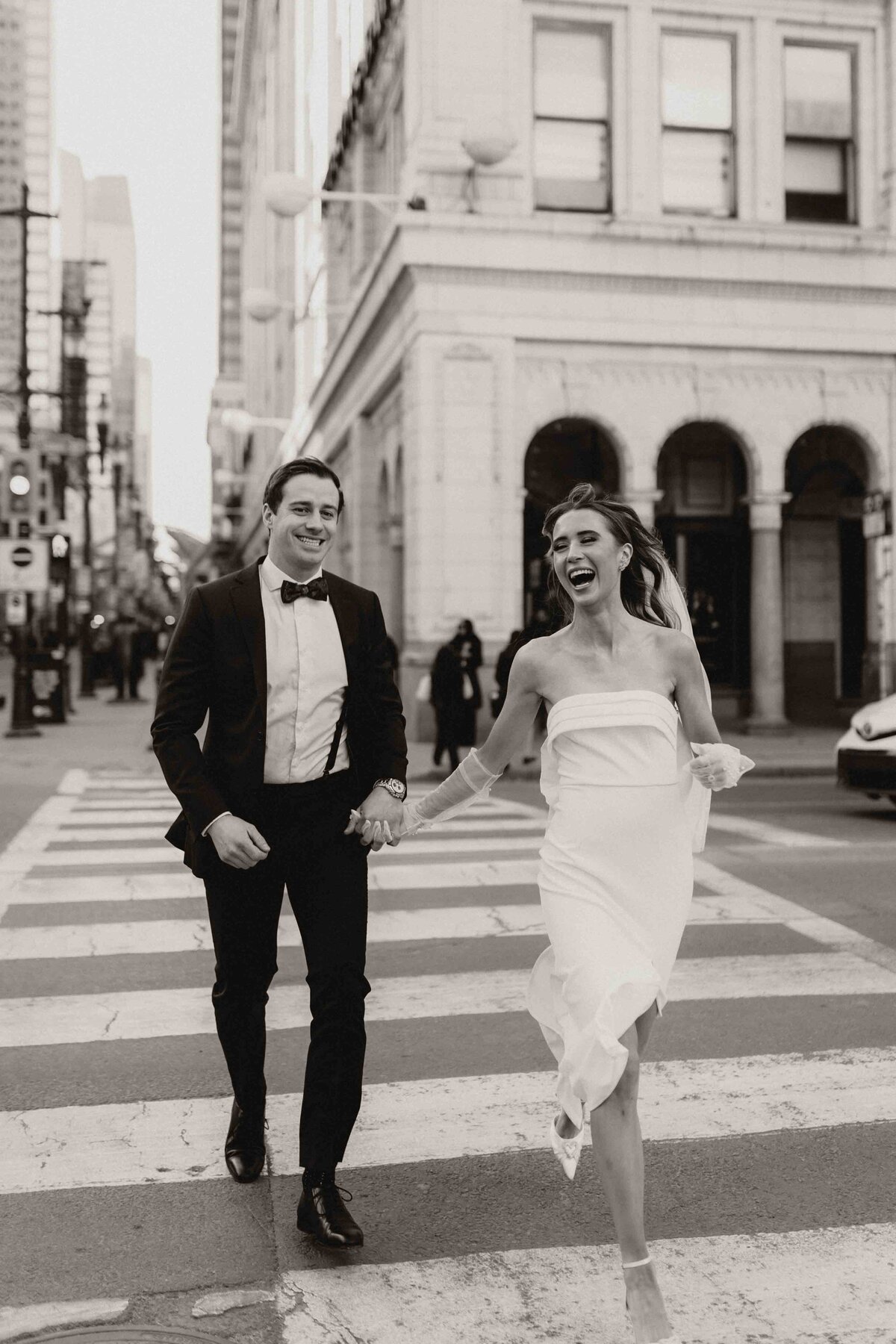 Black and white photo of a bride and groom running through the streets holding hands.
