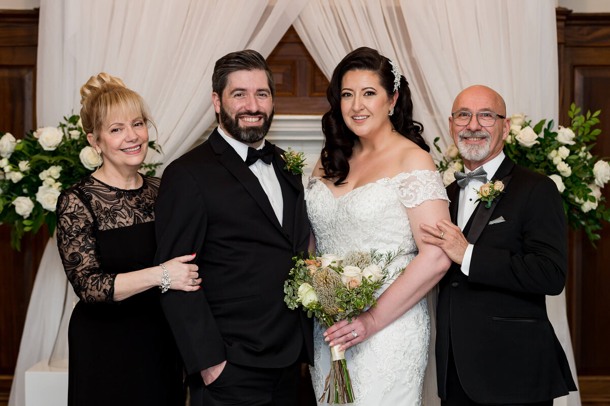 a groom in a black tux and a bride in a white dress stand together smiling with their parents in front of the Chateau Laurier fireplace in Ottawa