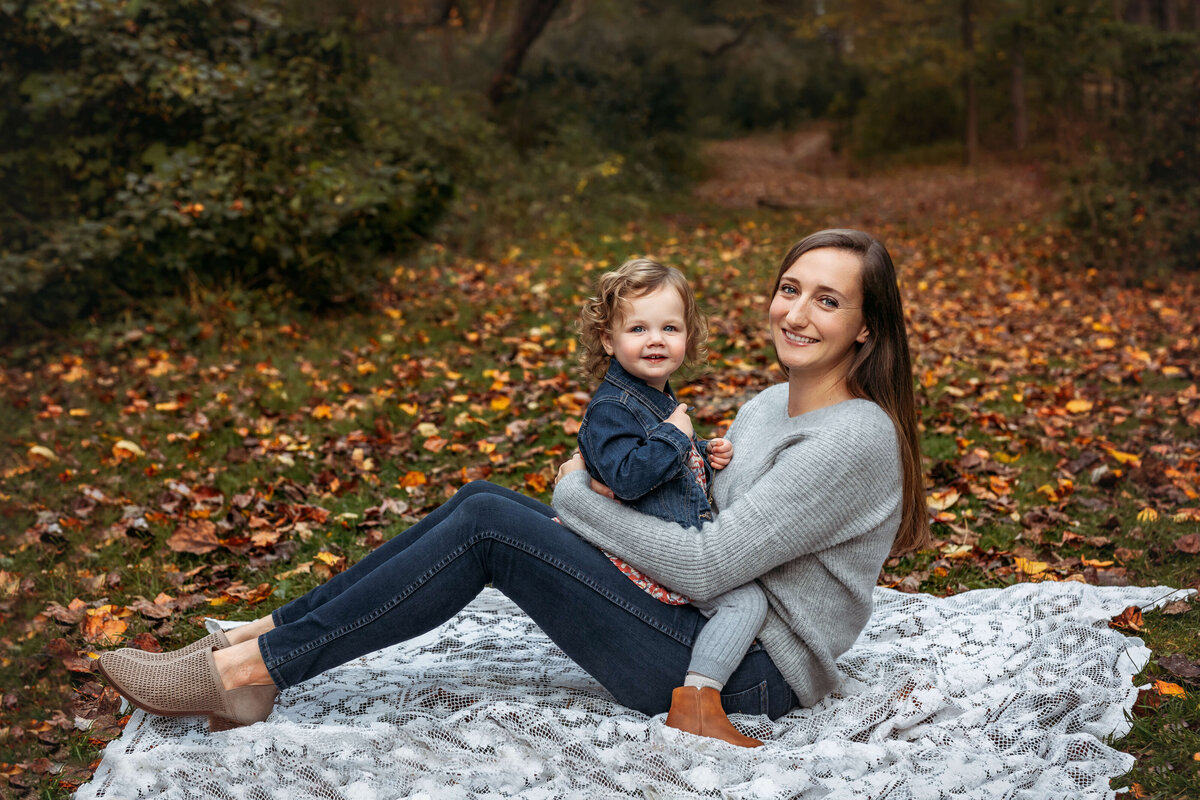 mom sitting to the side with her daughter on her lap smiling at the camera surrounded by fall leaves