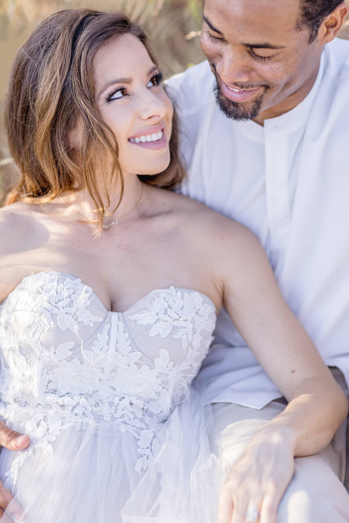 A couple sits closely, smiling at each other. The woman is wearing a strapless white lace gown, and the man is wearing a white shirt. They appear happy and relaxed in a natural outdoor setting.