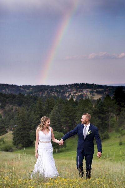 A bride and groom walk hand-in-hand toward the camera with a rainbow in the background.