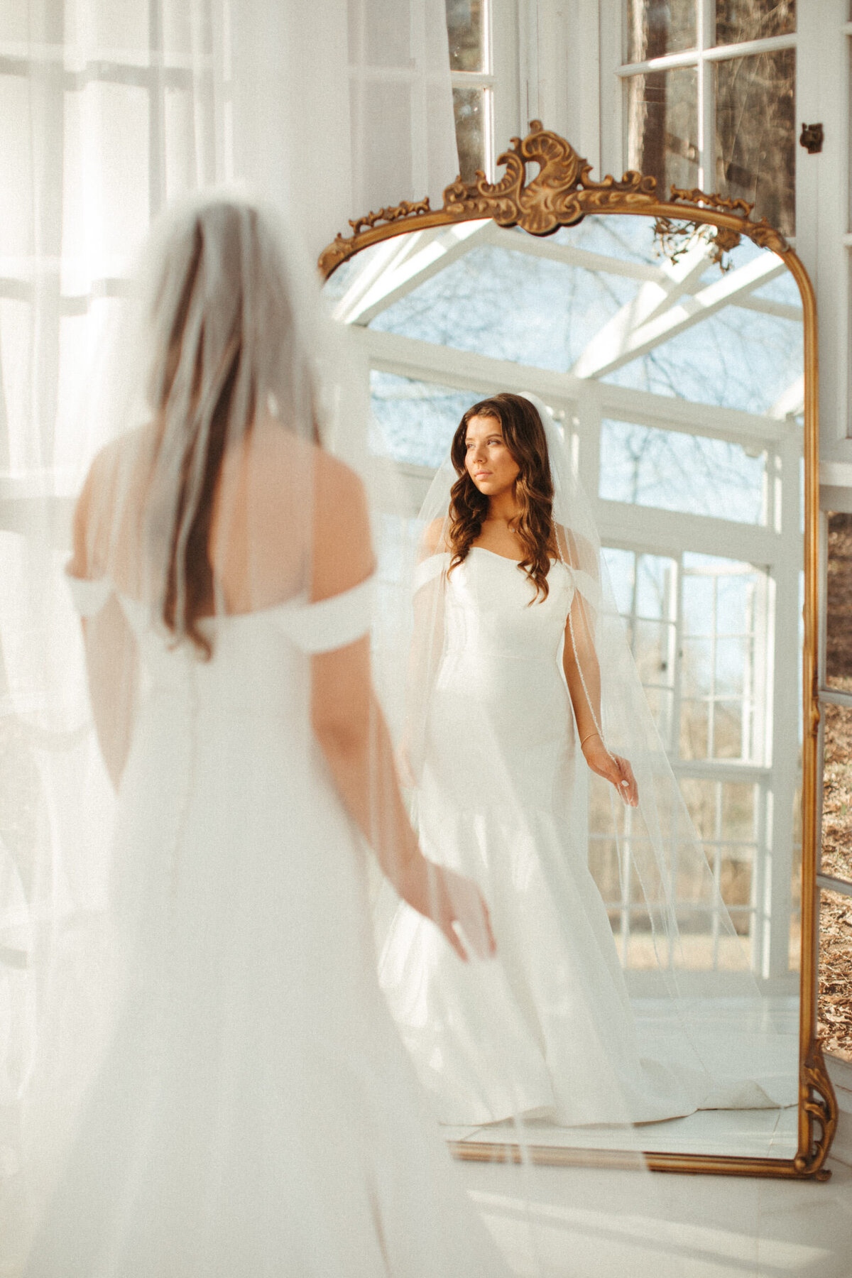 Classic bride with long veil standing in front of a full length mirror looking off