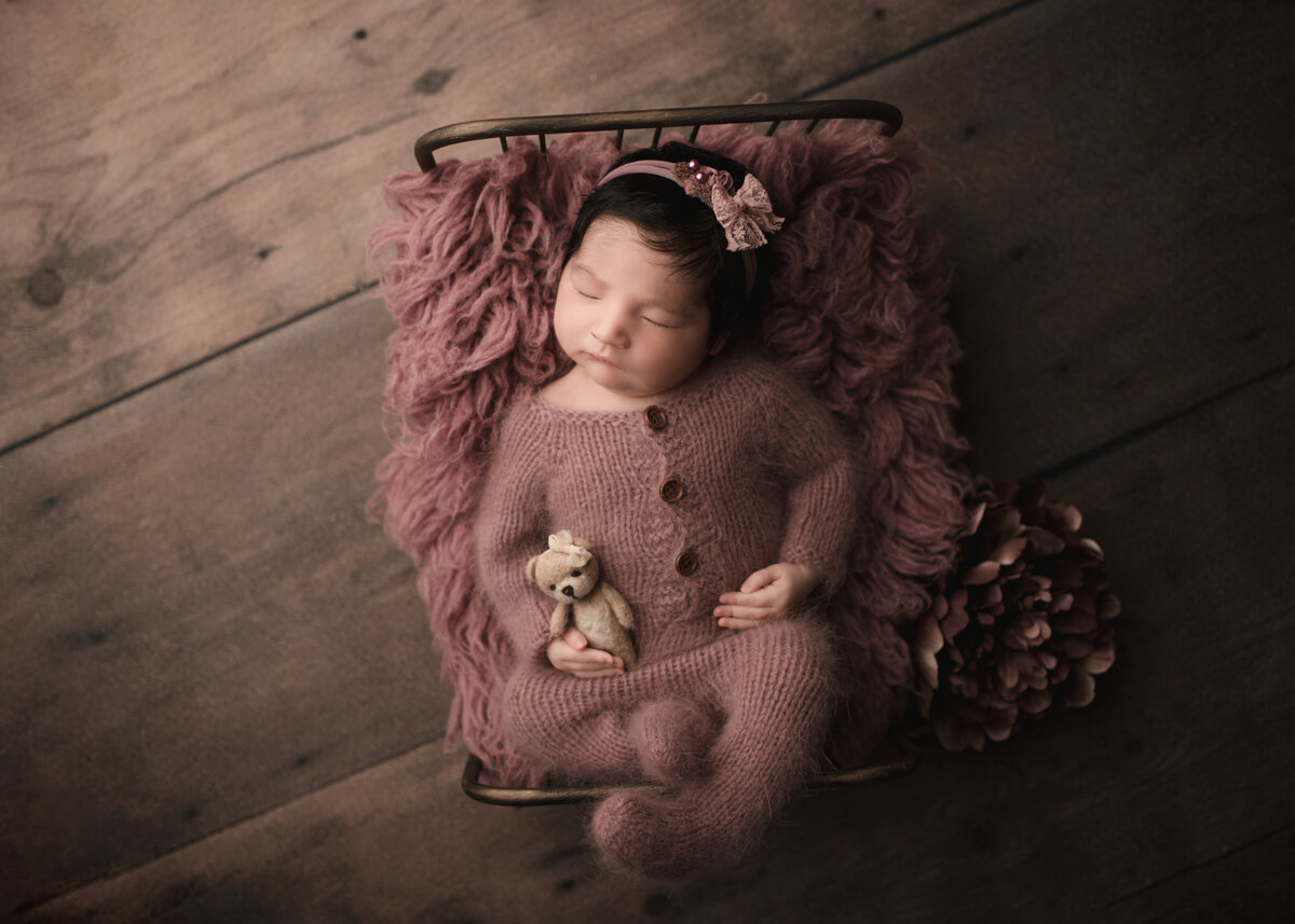 Aerial image of Riverside, CA newborn photoshoot. Baby girl sleeping on her back on a newborn prop bed. She is wearing a mauve knit romper. Her legs are folded over the foot of the bed and a tiny felt teddy is wrapped under one arm. Captured by Best Riverside, CA newborn photographer Bonny Lynn Photography.