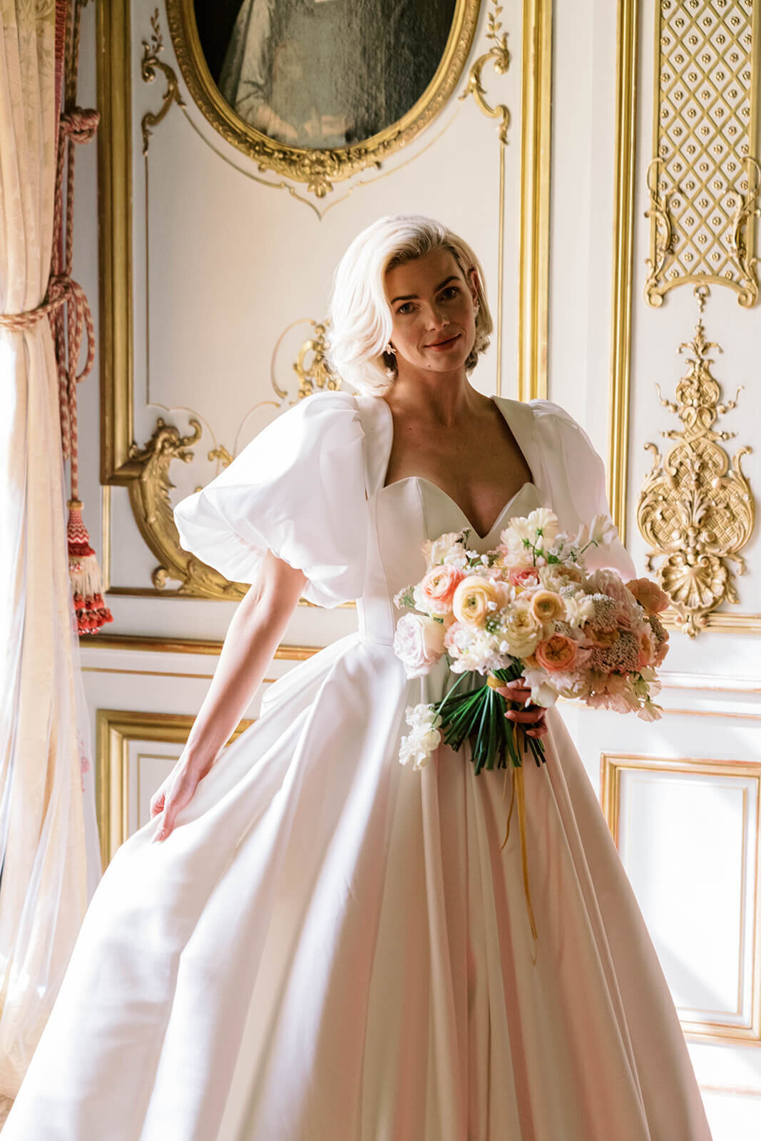 bride standing in blenheim palace wearing a luxury wedding dress with large puffed sleeves holding a peach wedding bouquet