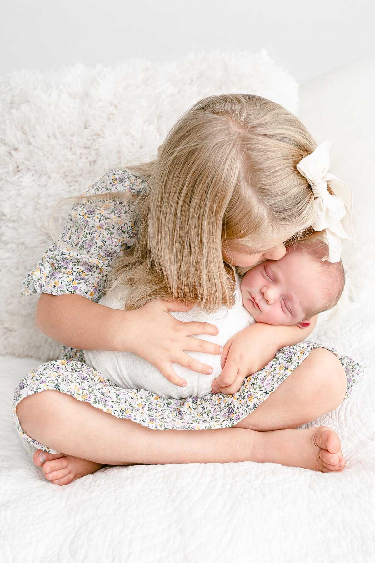 Little toddler girl sitting crossed-legged and holding her baby brother in her arms while sitting on a bed with neutral covers and pillows. She is kissing her baby brother on the temple with her blonde hair cascading down over him. Baby is sleeping.