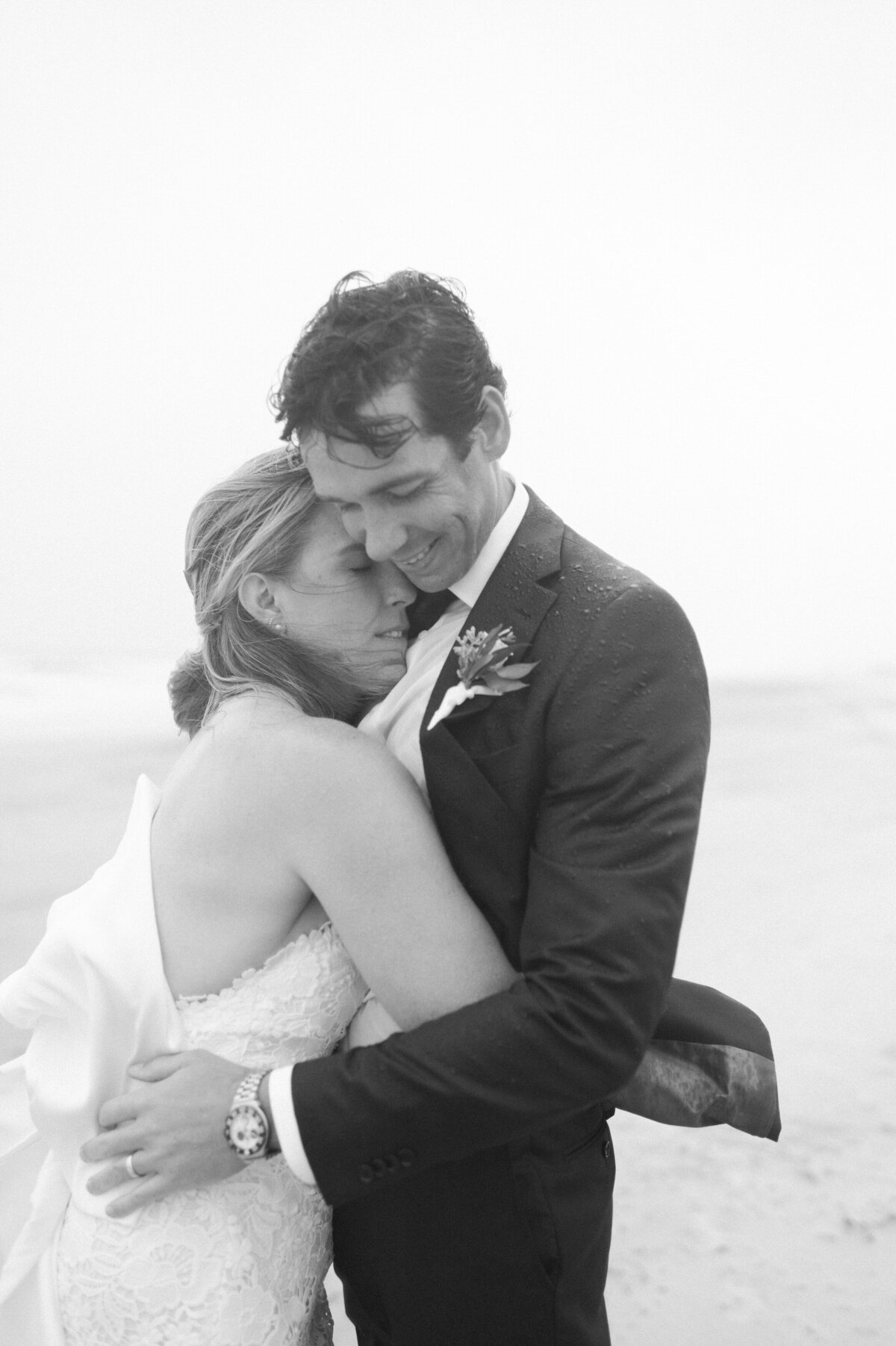 A bride rests her head on the groom’s chest as they hug in the rain