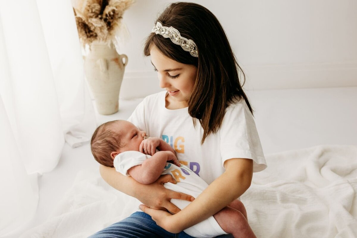 A sweet image of an older sister gently holding her newborn baby brother or sister, gazing at the infant with affection. A heartwarming sibling bond