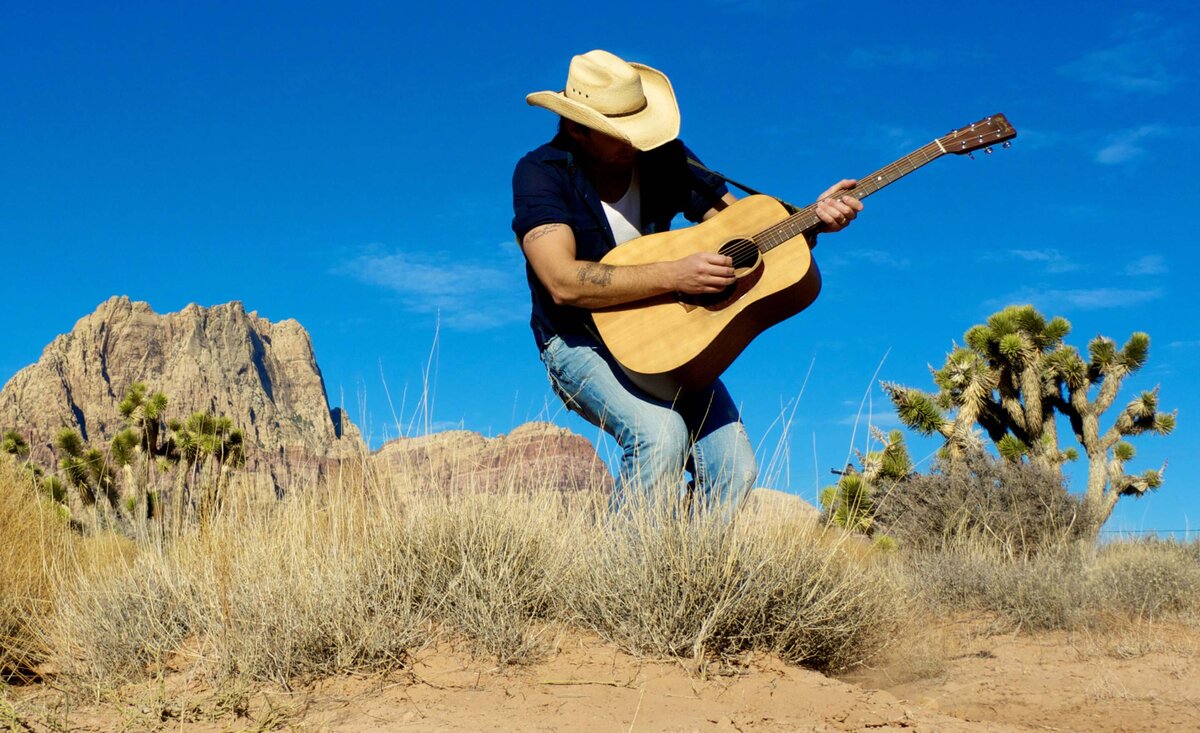 Male musician portrait Dean Brody wearing blue shirt blue jeans straw cowboy hat playing guitar amidst desert background