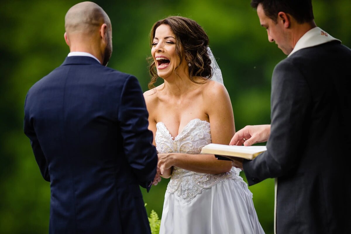 An exuberant bride in a detailed strapless gown laughs candidly during an outdoor wedding ceremony, with the officiant and groom smiling beside her.