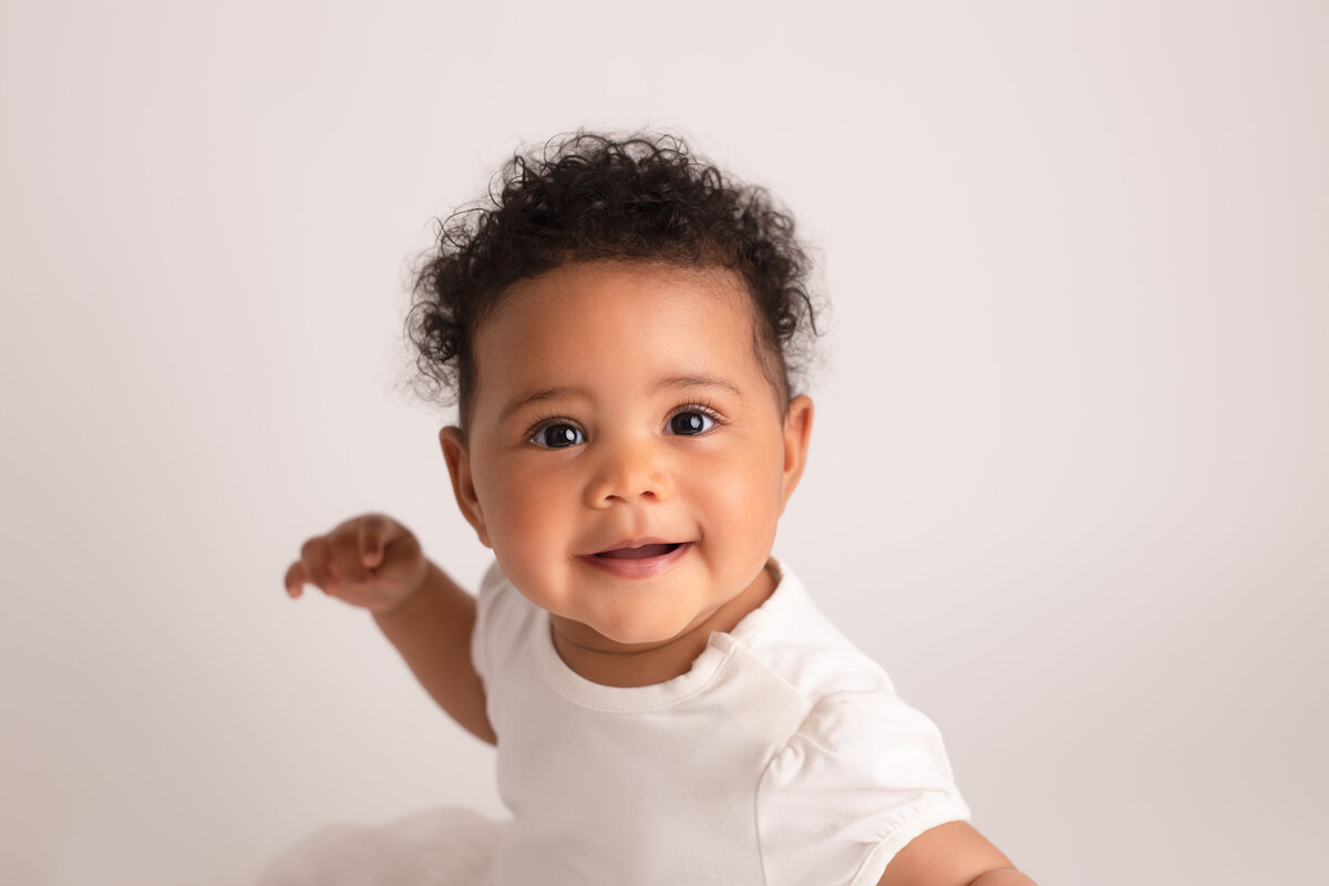 Happy six month old baby girl wearing a white dress on a simple white background.