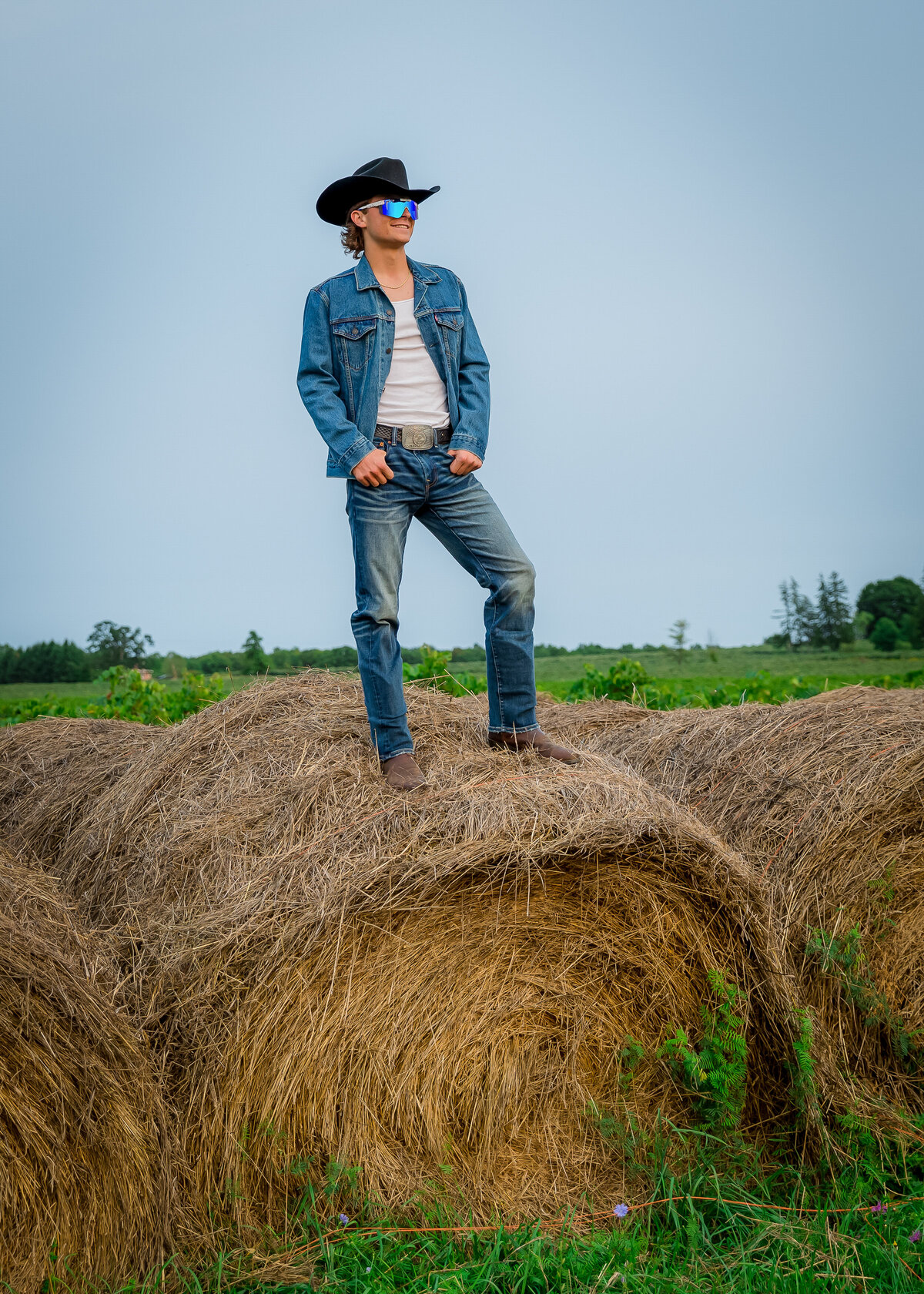 boy senior portraits cowboy farm on a hay bale wearing cowboy hat and Pit viper sunglasses in penn yan ny