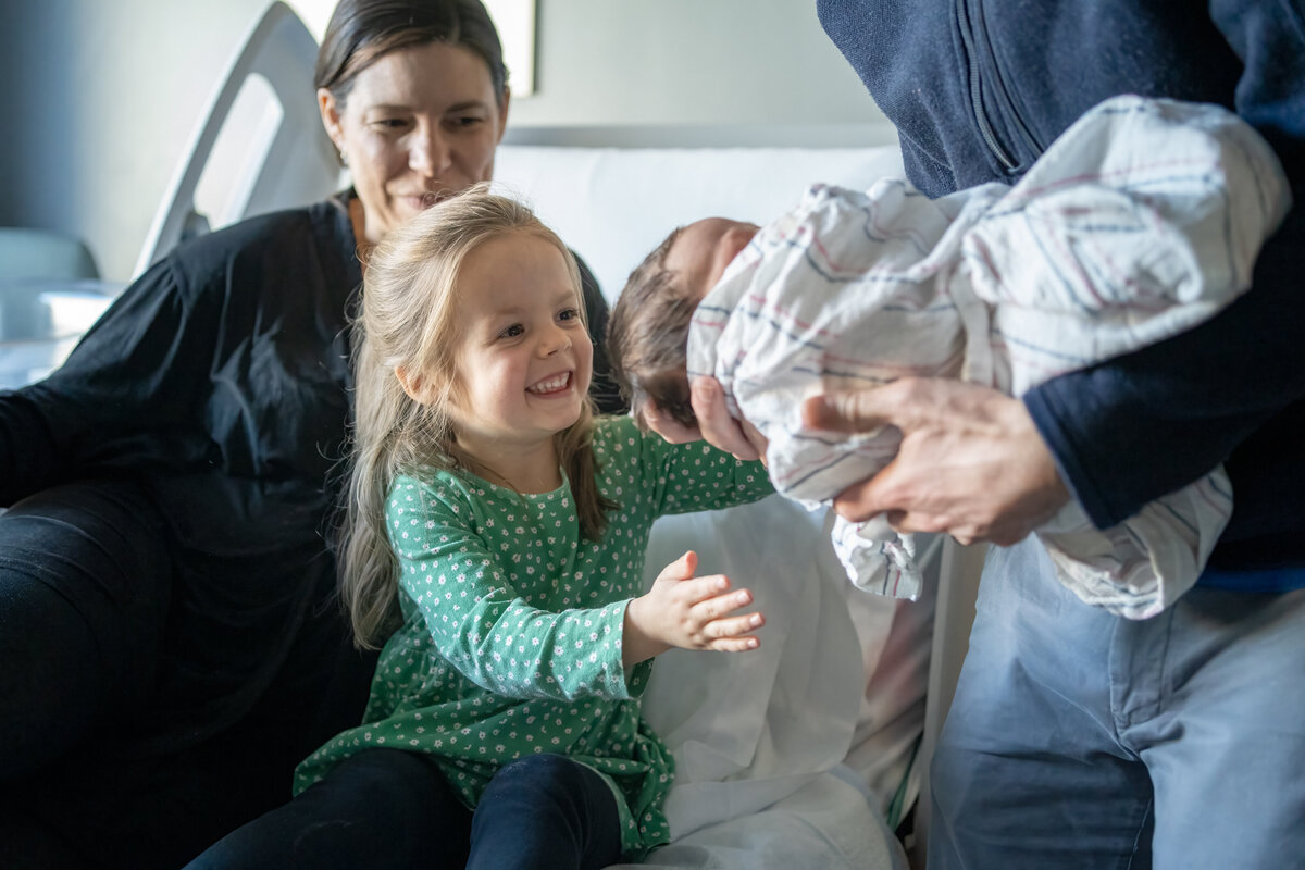 Toddler sitting on bed with mom reaching to hold her newborn baby brother