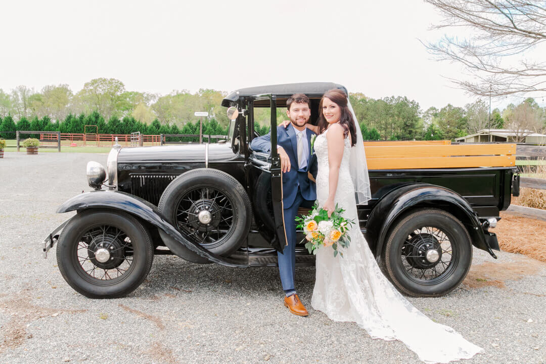 Groom sits inside a vintage car while his bride stands next to him.