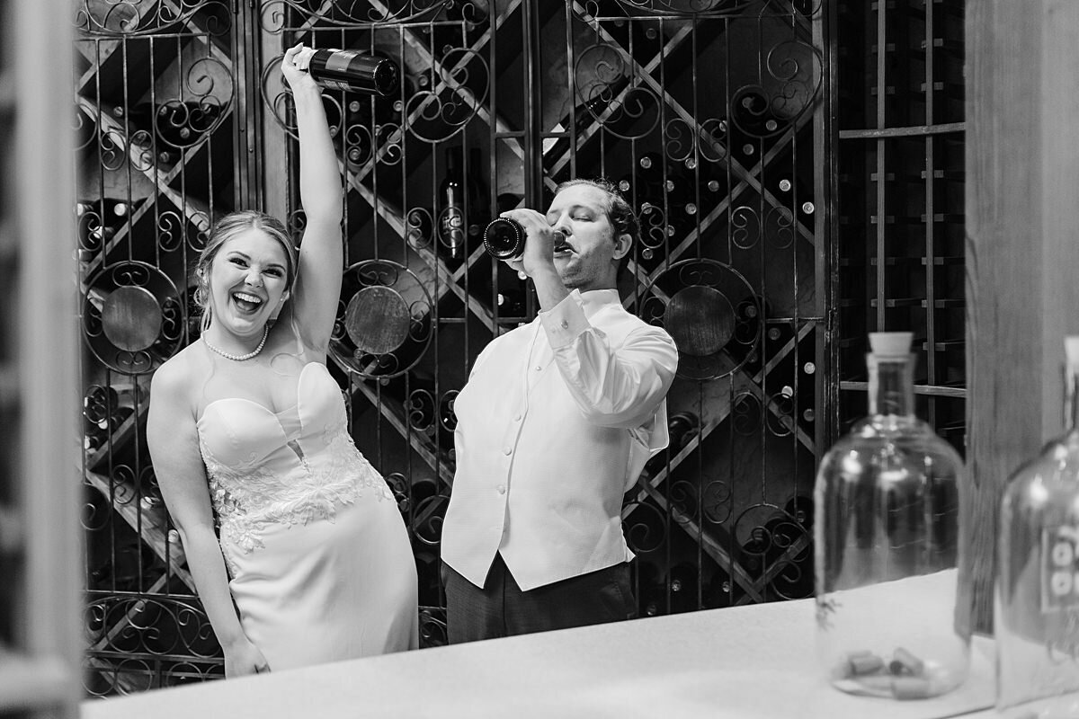 The bride, wearing a sleeveless fitted wedding dress, holds a bottle of wine up as she toasts her groom. The groom, wearing a white shirt and vest, stands in the wine cellar at The Estate at Cherokee Dock drinking a bottle of wine.