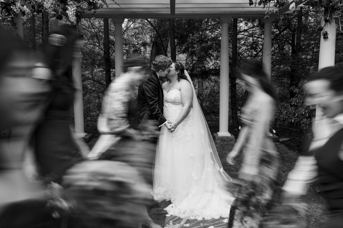 Brides kissing while bridal party passes by in a blur