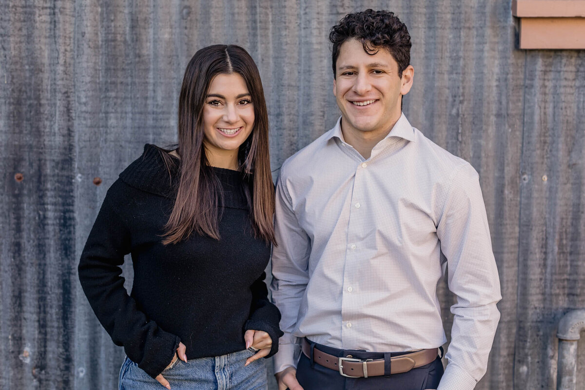 A young woman with long, brown hair wearing a black sweater with her hands in her pockets, smiling at the camera beside a young man with dark hair wearing a grey dress shirt also smiling at the camera in front of a grey metal wall.