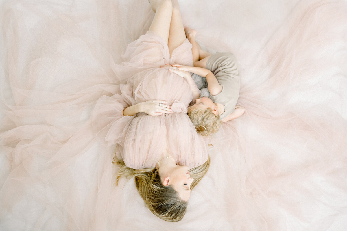A maternity session where the mother is photographed laying down on the ground dressed in a long tulle maternity gown while she is snuggling with her toddler son and holding her belly.