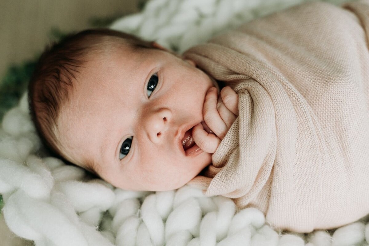 Awake newborn close-up – Close-up of a newborn baby swaddled in beige knit, awake with bright eyes open and tiny fingers near the mouth, lying on a chunky white blanket.