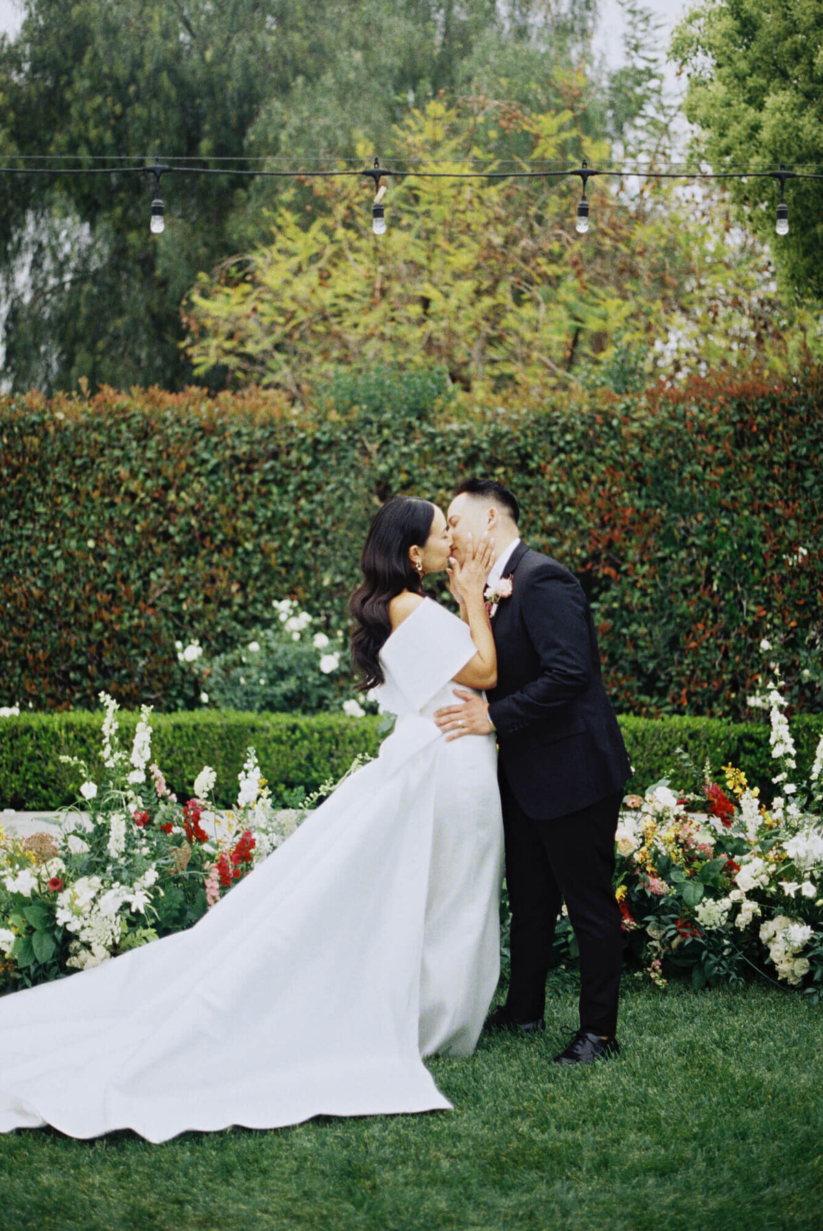 A bride in a white gown and a groom in a black suit kiss in a lush garden, surrounded by colorful flowers and greenery.
