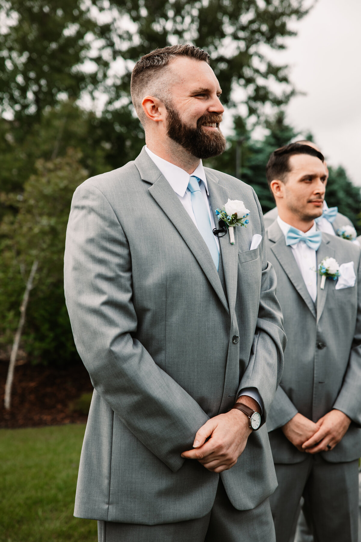 A happy groom in a grey suit waits for his bride to walk down the aisle of their outdoor wedding