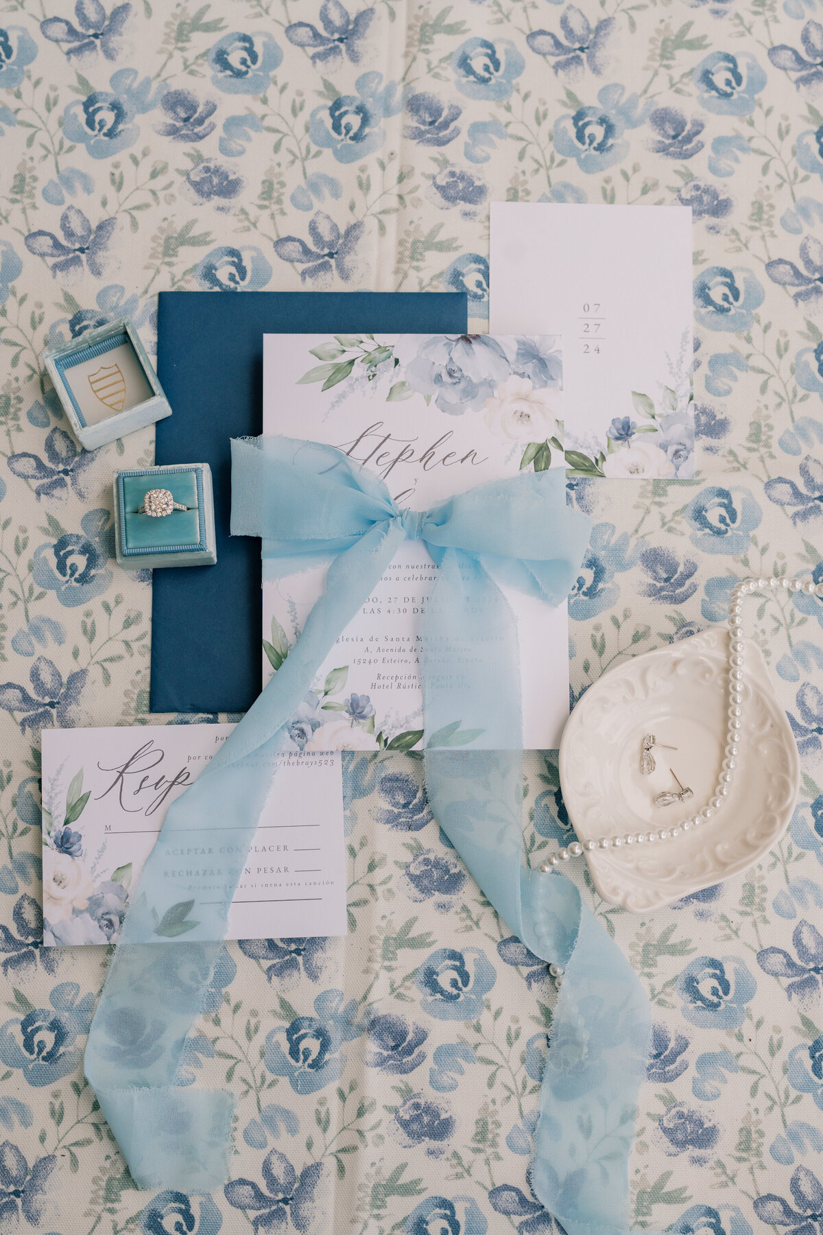 Weddings in Spain | A white wedding invitation with blue ribbon and a navy ribbon on a blue floral background. A blue ring box holding a silver engagement ring and a white vintage dish holding silver earrings and a pearl necklace surround the invitation suite.