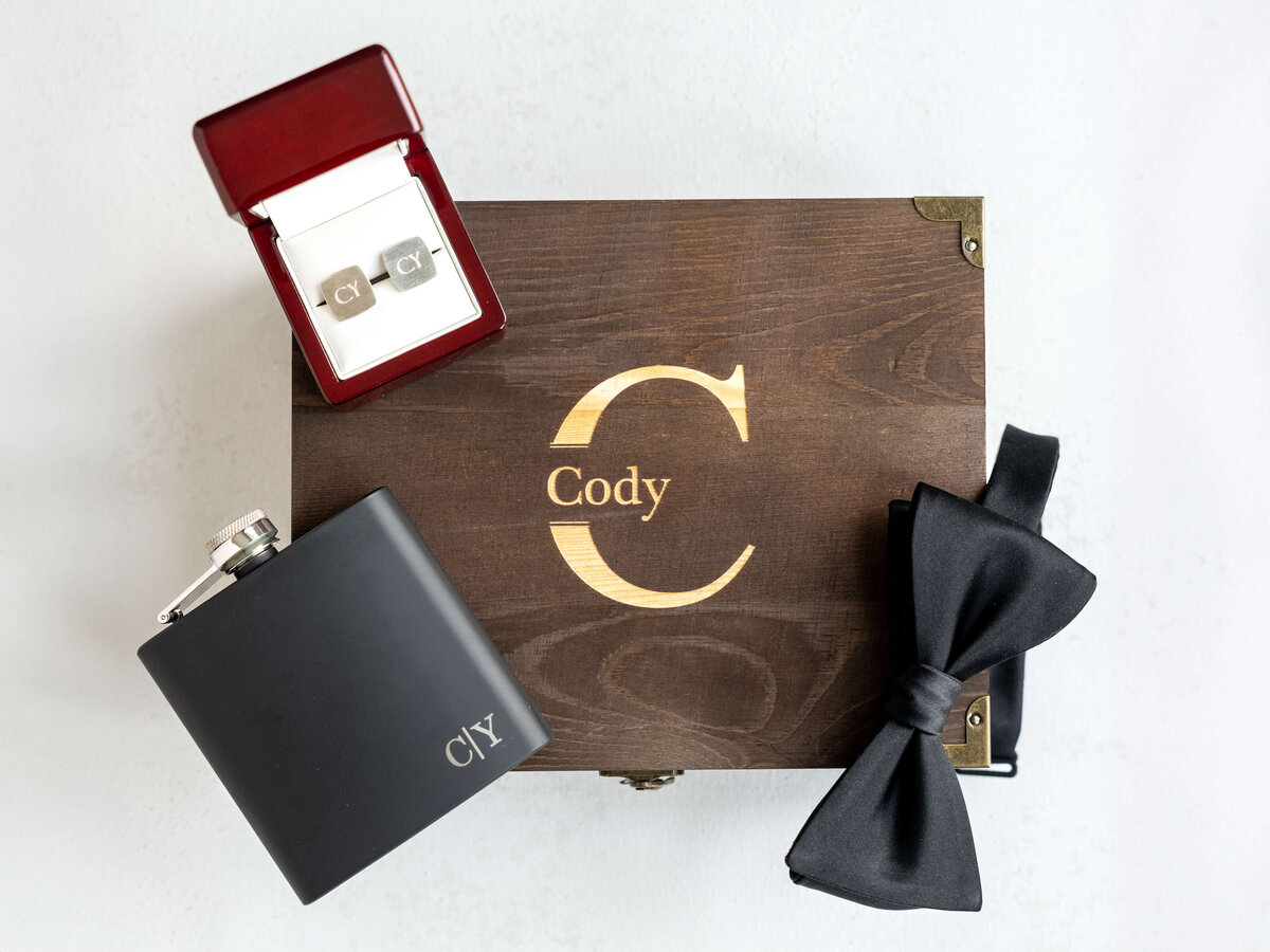 A stylish gift set for men featuring wooden box with engraved Cody, silver cufflinks in a red box, a black hip flask, and a black bow tie, all arranged on a light background.