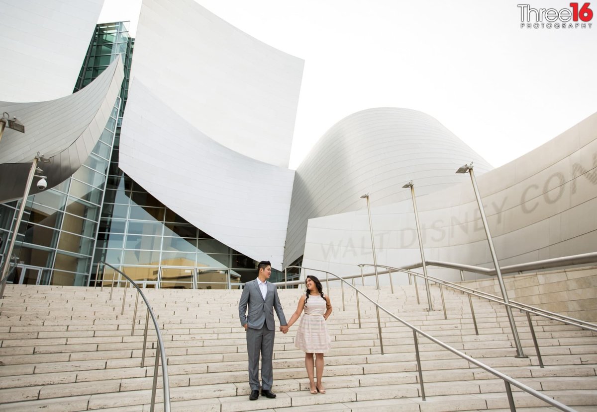 Engaged couple stand on the stairs of the Walt Disney Concert Hall holding hands during an engagement session