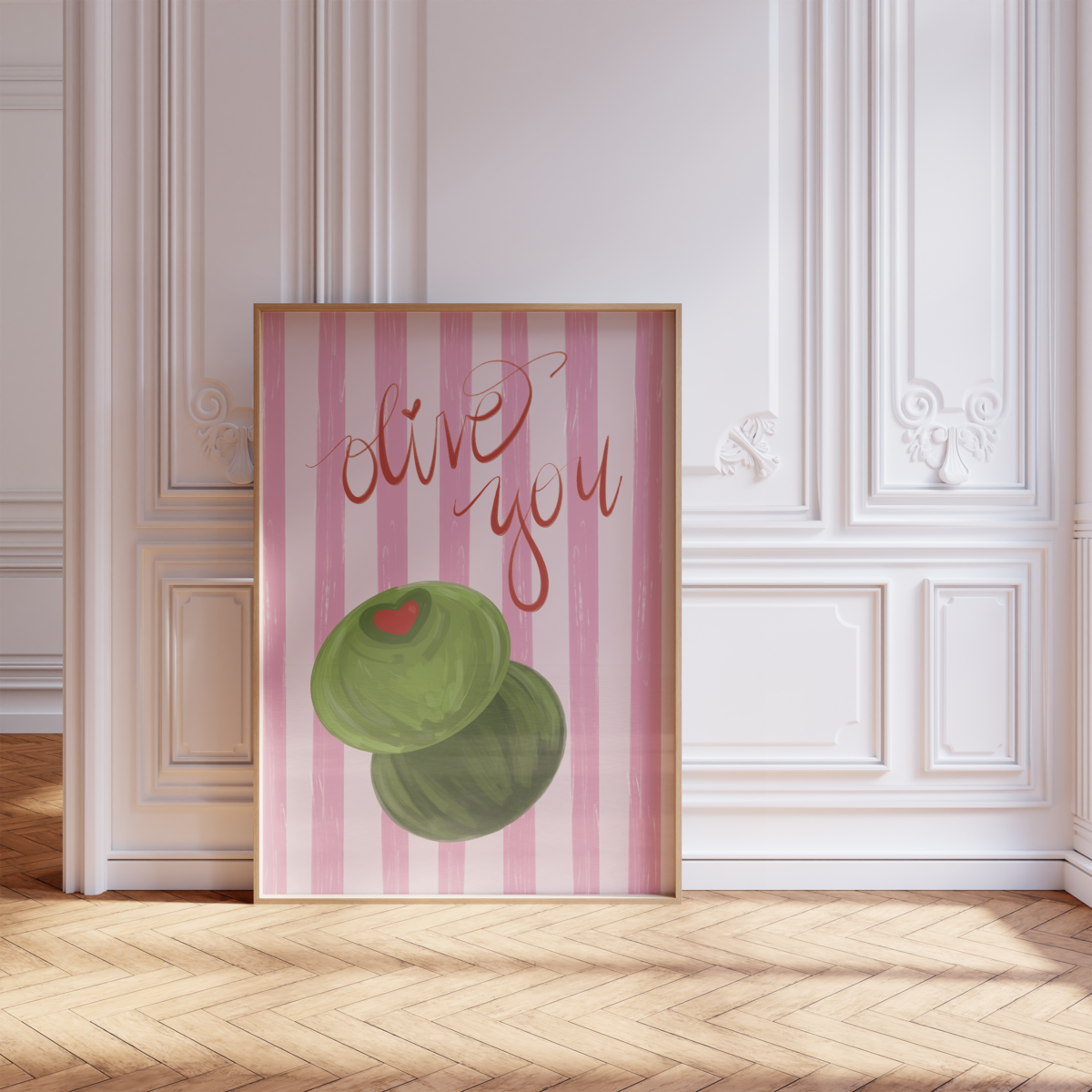 A painting by Sweet Pea Ink Creative features two green olives with a red heart, "olive you" text, and pink stripes. It hangs on a white wall in an artist studio, alongside a stool and paint-stained wall—perfect brand design inspiration.