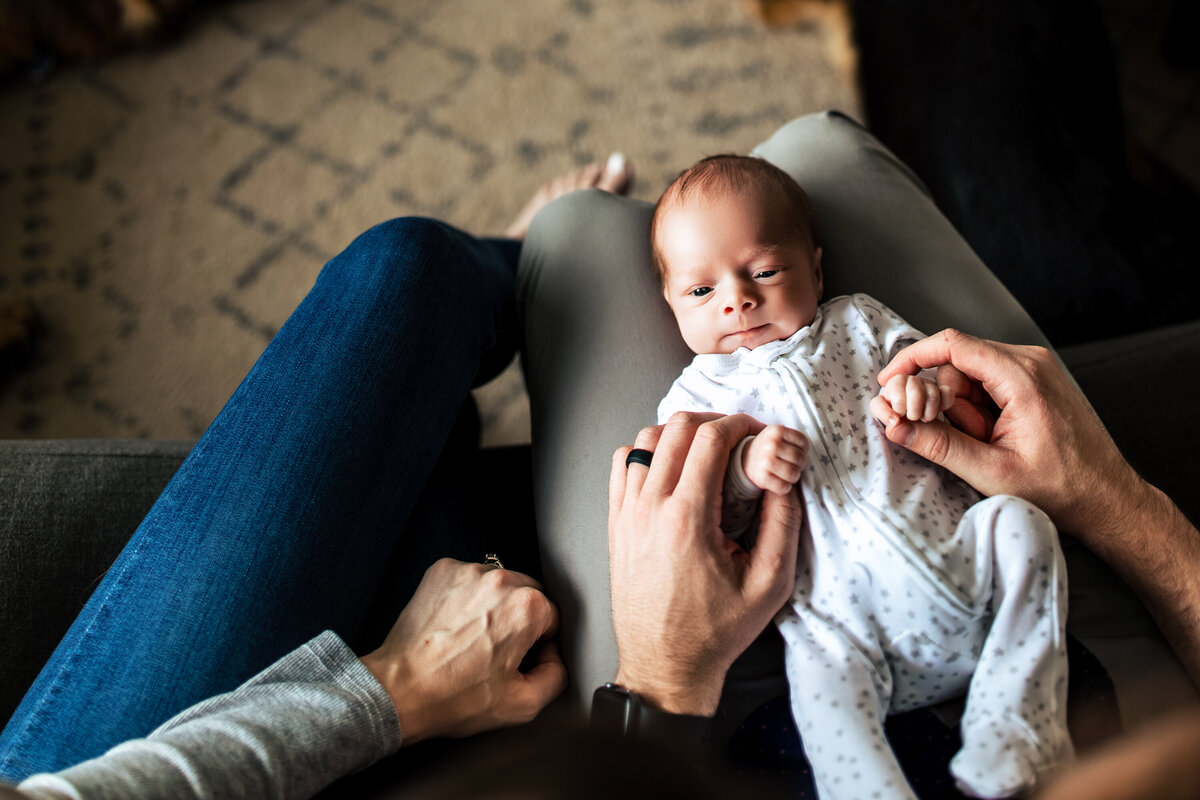 An in-home newborn Child Photo shoot a baby has his hands held by his father as he stares up into his mother's eyes for a portrait