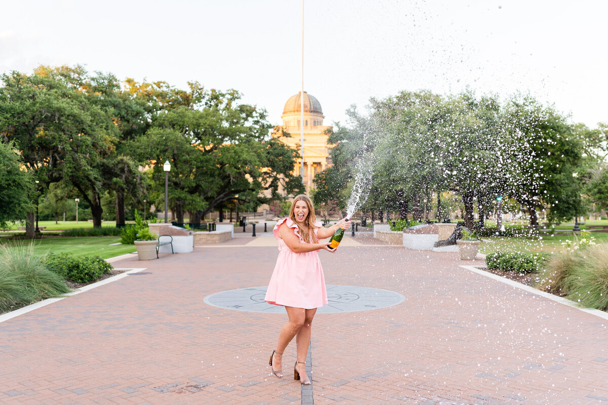 Texas A&M senior girl spraying champagne and laughing while wearing pink dress in front of Academic Building in Military Plaza