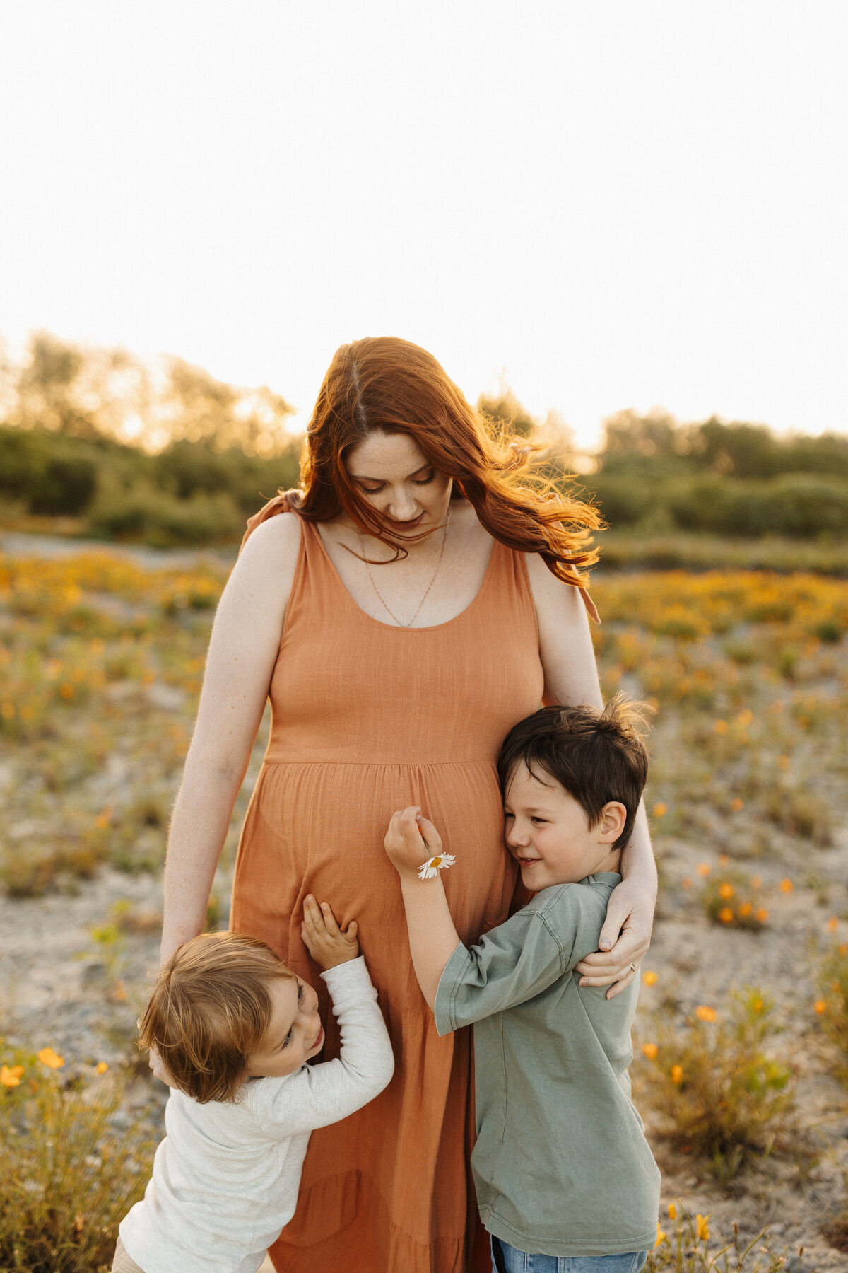 A mother with her two young children at sunset while the wind is blowing her hair around and she is directed by Christchurch photographer Lucy Jane Photography