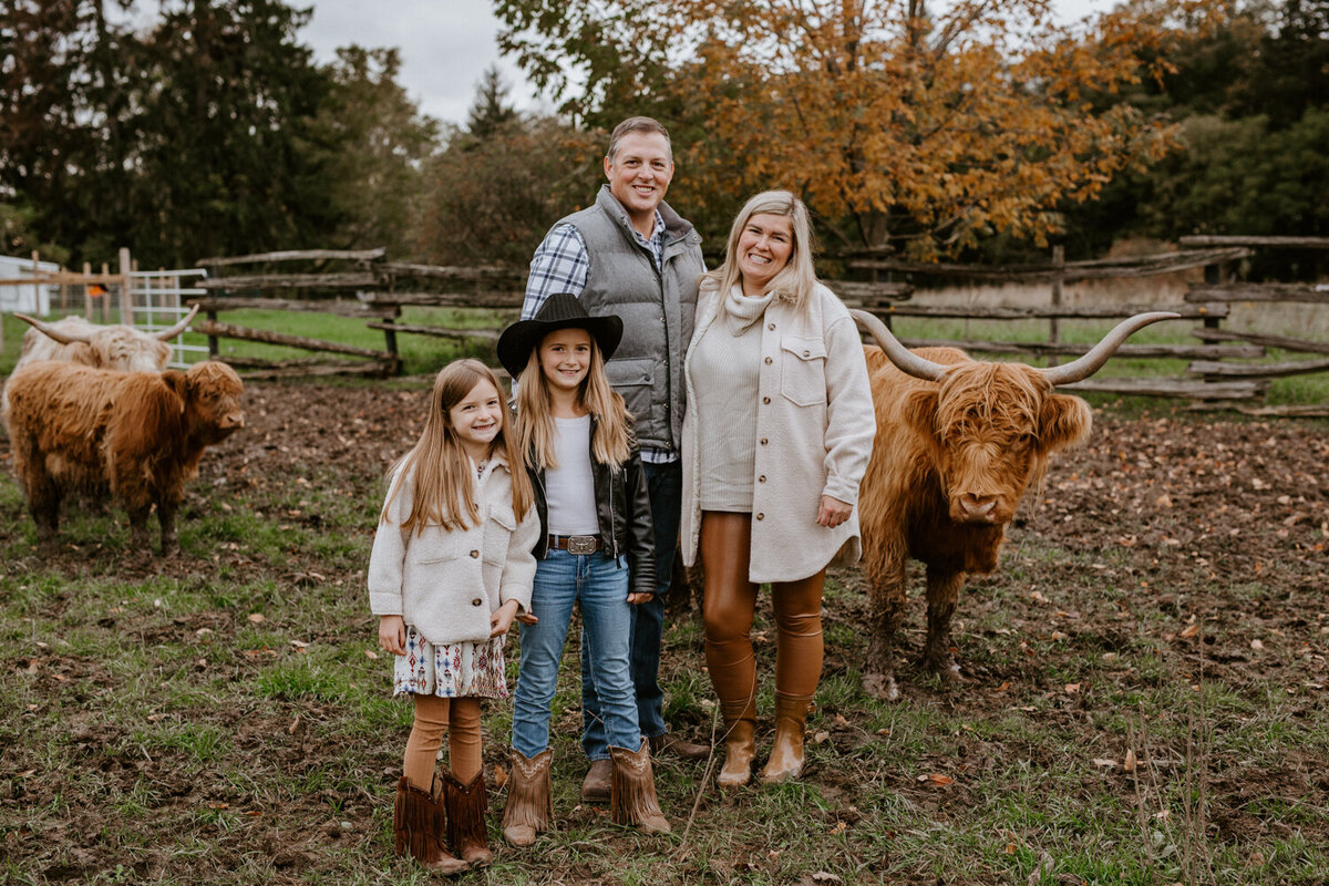 Family photoshoot with highland cattle outside of London, Ontario. Mom, Dad, and two tween daughters are standing in the pasture smiling at the camera. There is a highland cattle beside them looking at the camera.