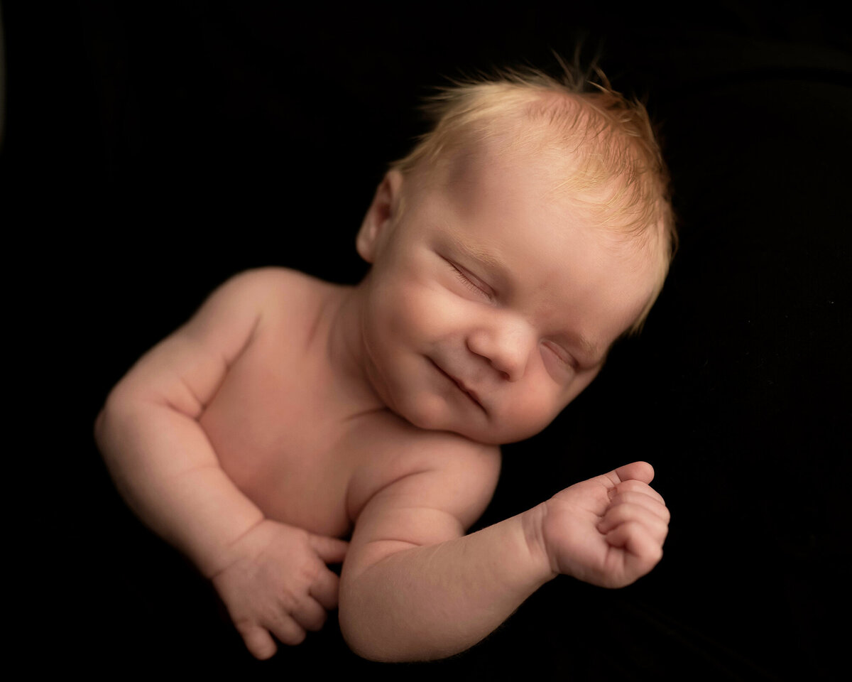 A newborn baby smiles in its sleep while on a black bed in the studio of a Pella Newborn Photographer