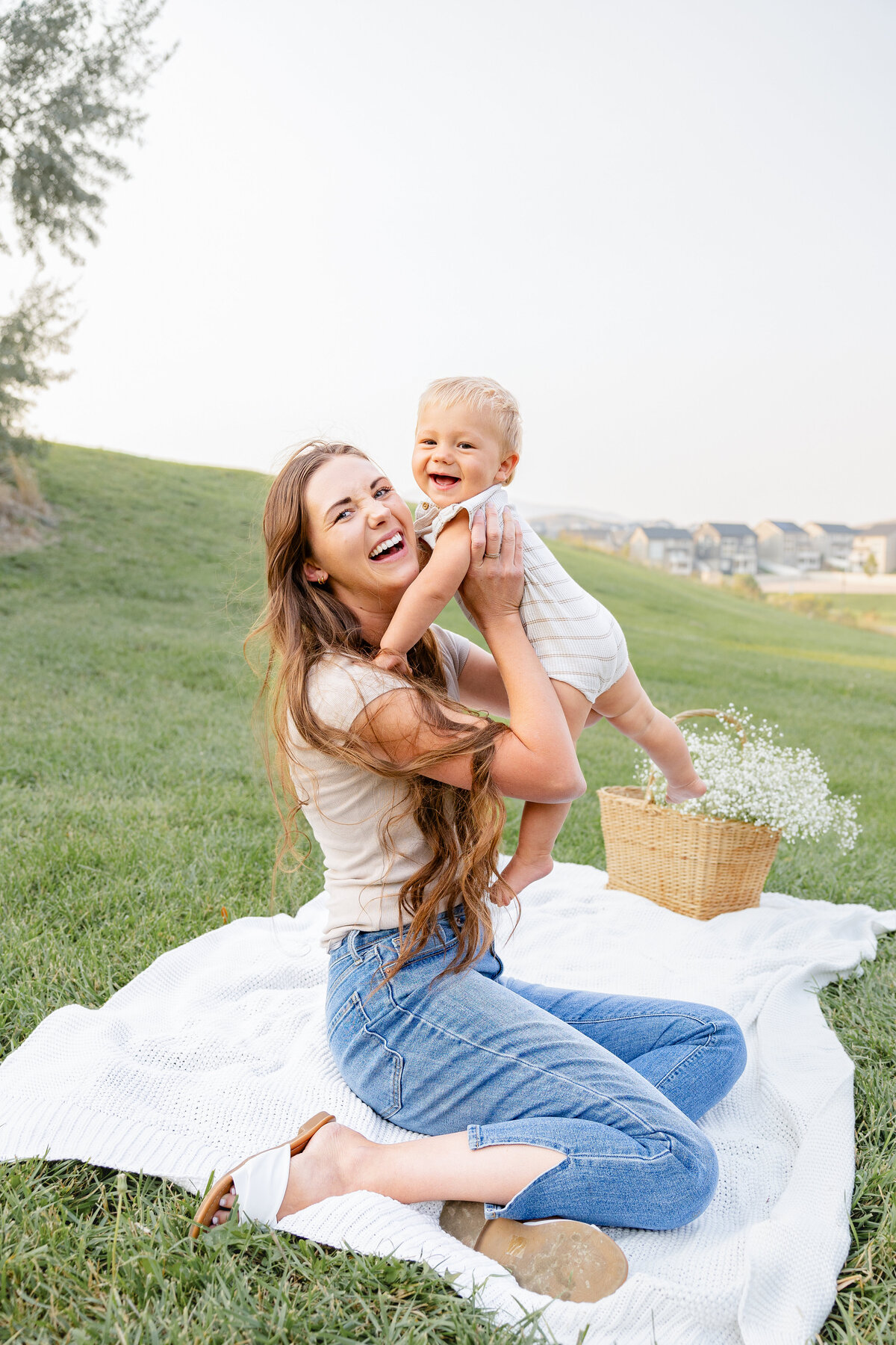 A mother lifts her baby into the air, both laughing joyfully in a serene outdoor setting. This warm and playful moment was captured by Cassie Lee Photography in Eagle Mountain, Utah.