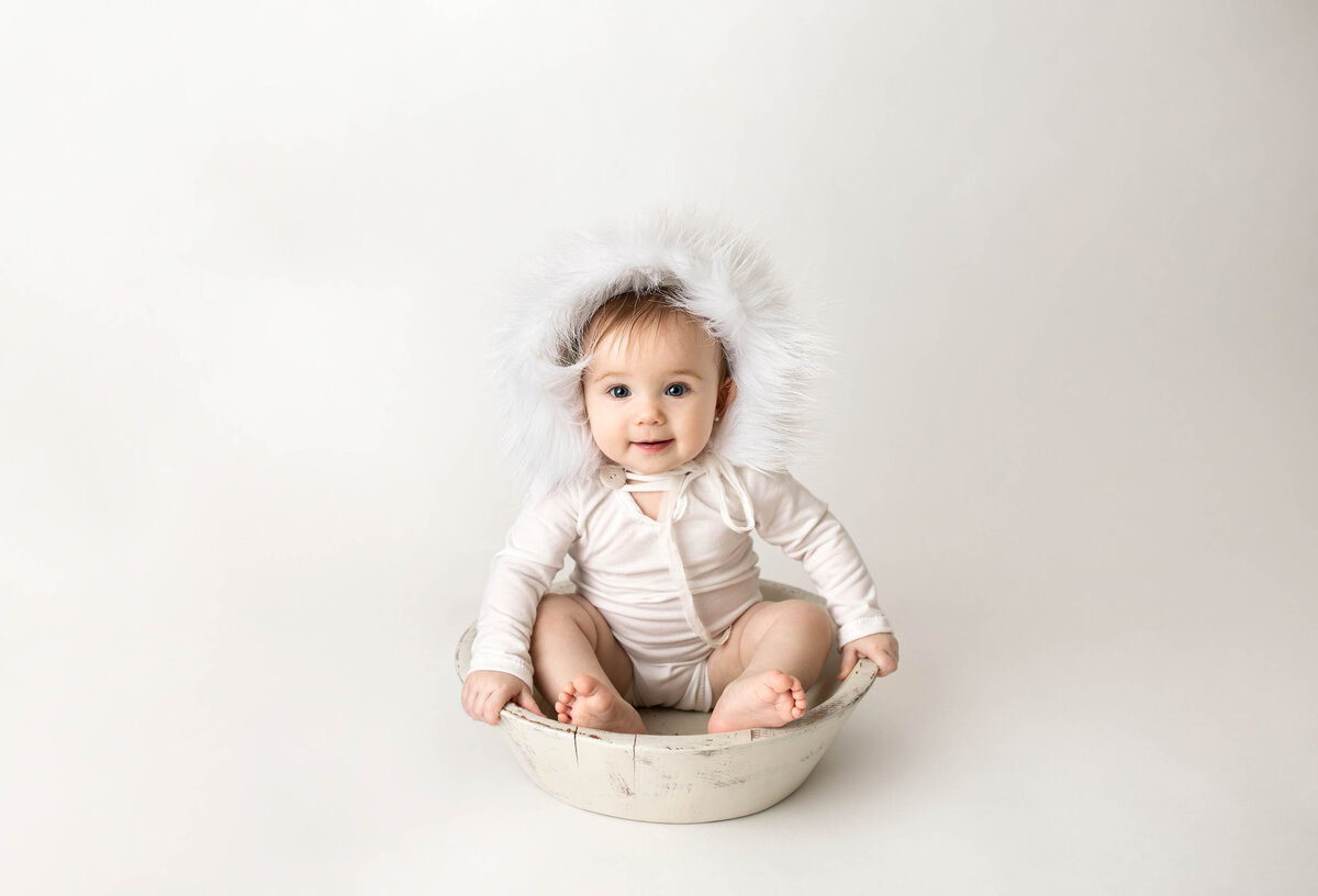 Baby sitting in white knit onesie and faux fur trimmed bonnet sitting on white backdrop in London, Ontario photography studio. Baby is smiling and looking directly at the camera.