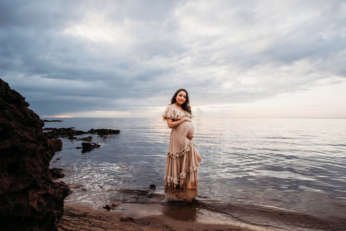 Pregnant woman stands in the water with stromy skies behind her at Melbourne maternity photography shoot