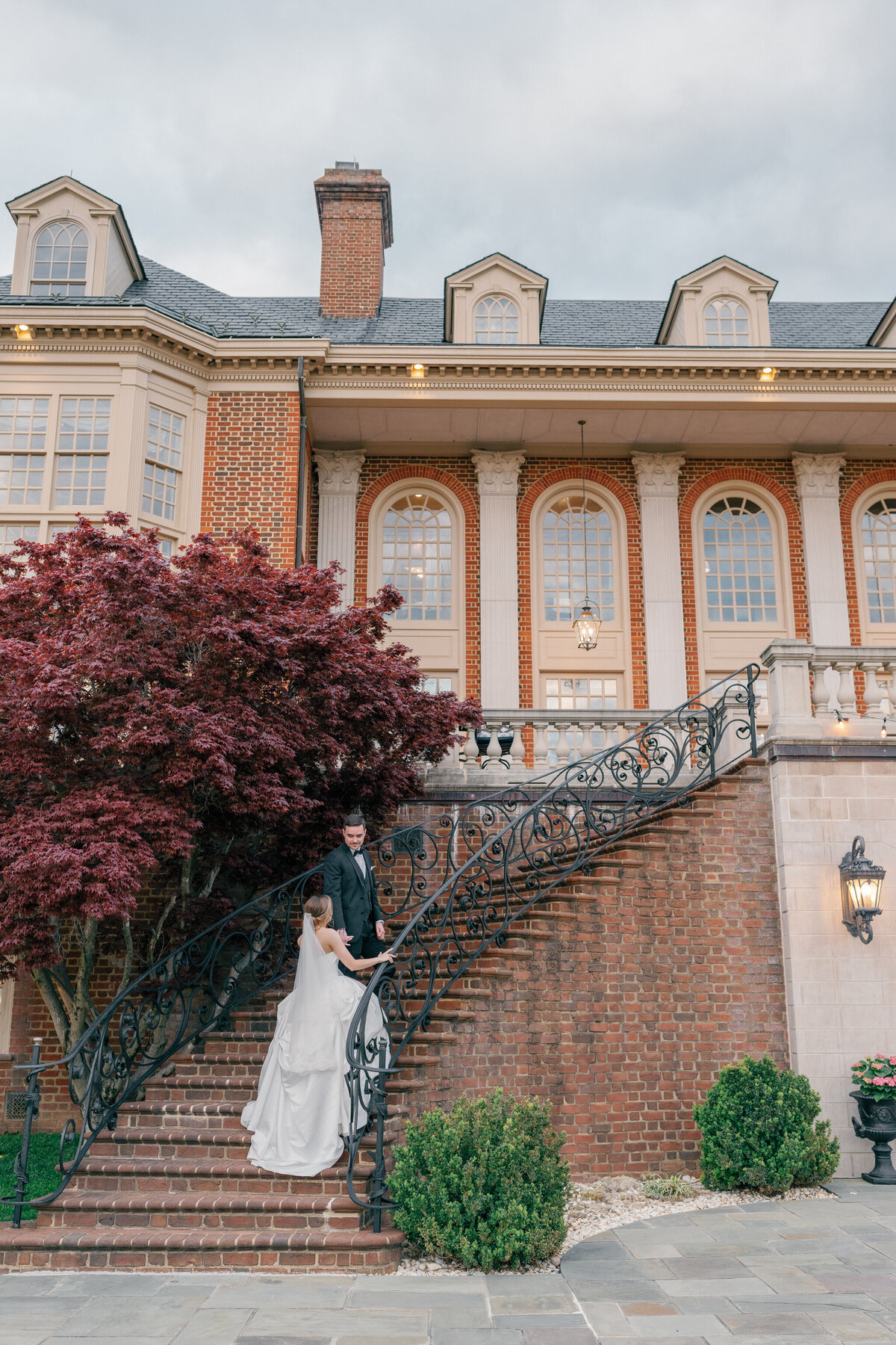 A bride in a flowing white gown ascends an ornate wrought-iron staircase, reaching for her groom’s hand at The Estate at River Run. The stately brick mansion with grand arched windows and lush landscaping provides a timeless, romantic backdrop. Captured by Caitlin Wilcox Photography, a Richmond, VA film wedding photographer.