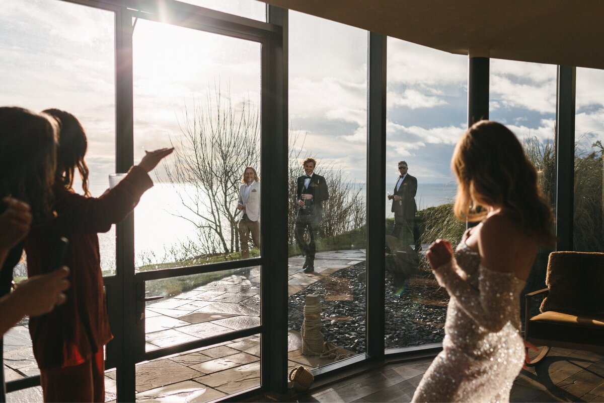 Groom first look at stunning bride through big glasswindows at the secret suit at Gara Rock 