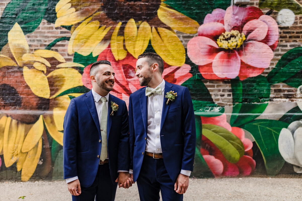 two grooms in blue suites hold hands in front of flower mural