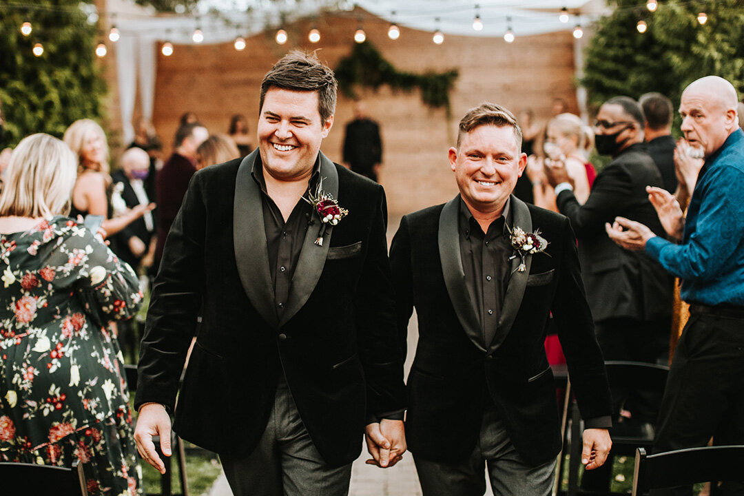 Two grooms wearing black tuxedos walk down the aisle holding hands at their wedding.