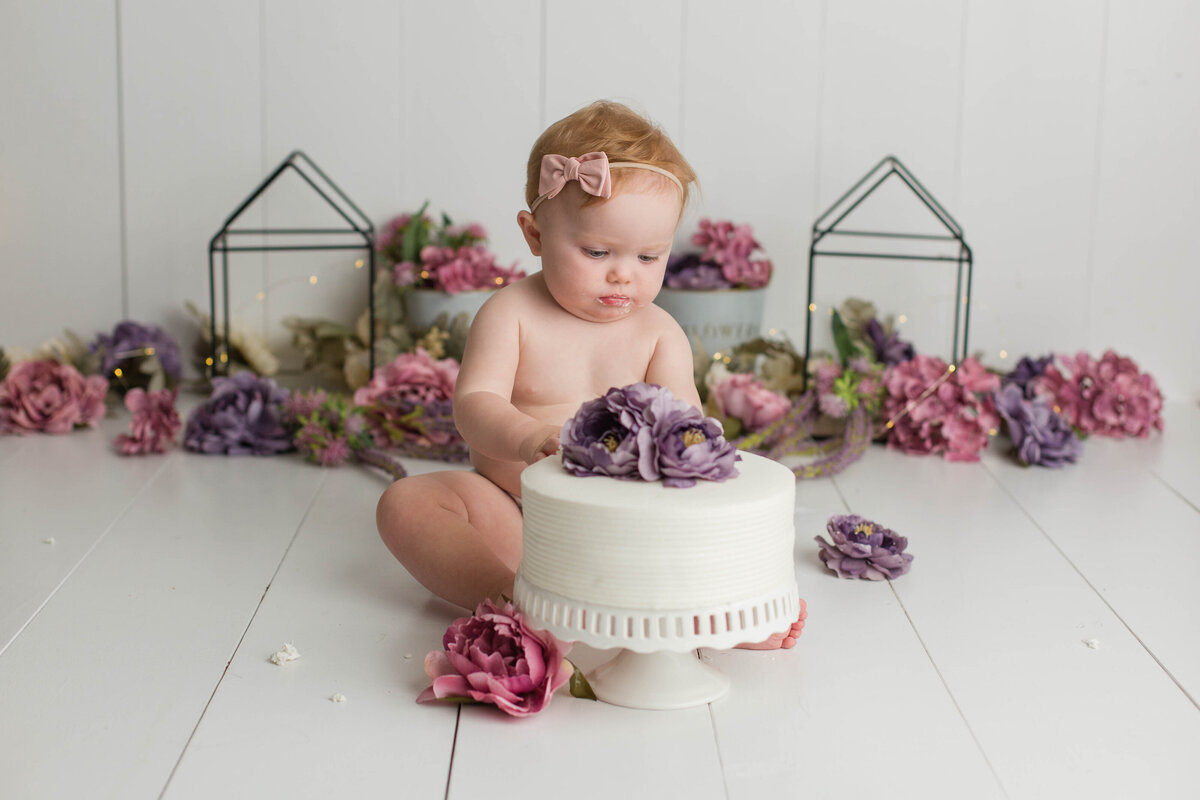 Baby intently looking at a cake decorated with purple flowers, pondering the next bite.