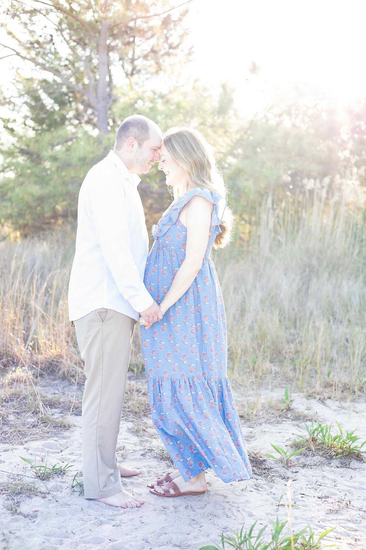 A pregnant woman and her husband stand facing each other holding hands in a coastal area by Mary Eleanor Maternity Photographer Virginia Beach