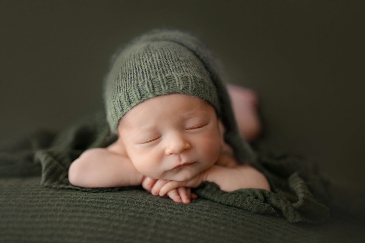 newborn baby boy laying on his stomach with his head on his hands wearing a green sleepy cap on a green backdrop  at his newborn photography session with a photographer at a arlington va photo studio