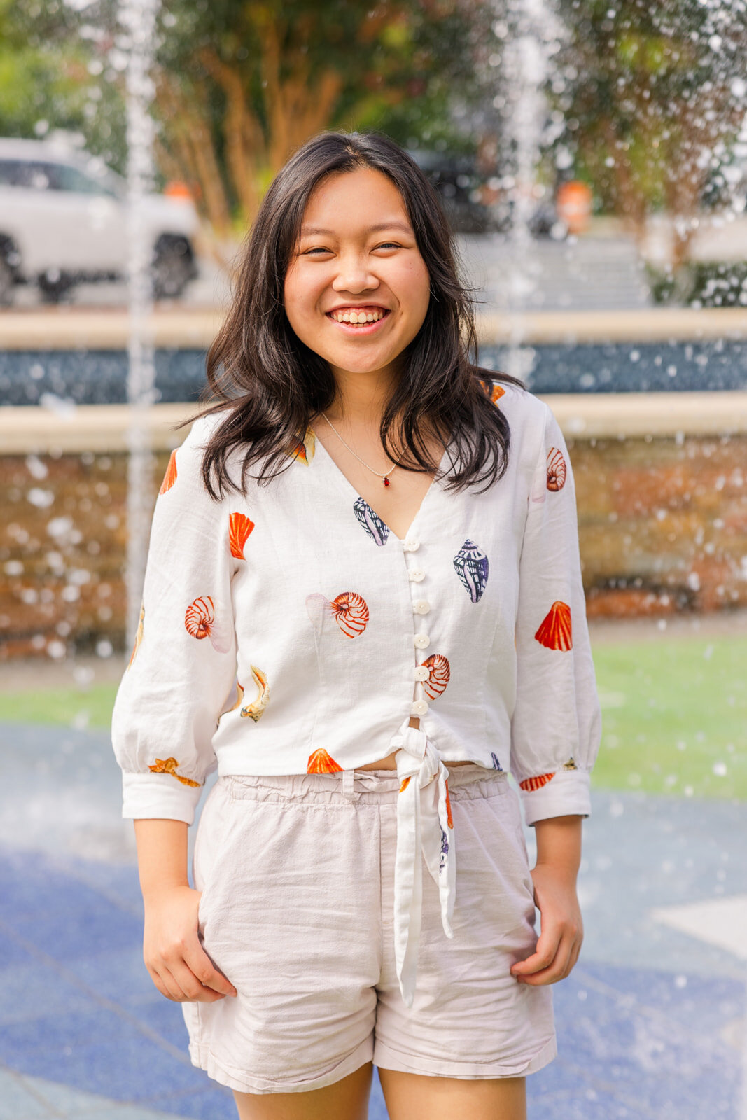 teenager girl in shorst and blouse with patterns, hands in pockets laughing at the camera with the fountain in the background in Sandy Spring during senior portraits session